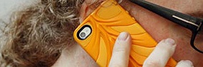 Eine Frau mit Locken und Brille hält ein orangefarbenes Smartphone an ihr Ohr