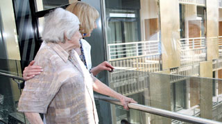 Pflegerin und ältere Dame mit Rollator stehen an einem Fenster