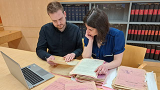 Eine Frau und ein Mann sitzen an einem Schreibtisch und sichten Archiv-Dokumente
