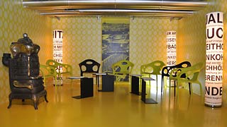 Gelb-grüner Ausstellungsraum mit Stühlen und einem Ofen