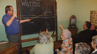Gruppe älterer Menschen in einer historischen Schulklasse; ein Mann zeigt mit einem Stock auf die Tafel