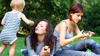 2 junge Frauen sitzen im Gras, neben ihnen spielt ein Baby
