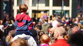 Viele Menschen auf einer Straße in der Stadt, ein Kind sitzt auf den Schultern eines Erwachsenen.