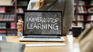Eine Frau, von der nur der Torso im Hintergrund sichtbar ist, sitzt hinter einem Laptop, auf dessen Bildschirm "(N)ever (S)top Learning" steht.