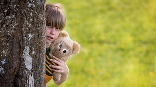 Kleines Mdchen mit Teddybr sieht hinter einem Baum hervor.
