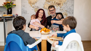 Um einen Tisch sitzen eine Frau und ein Mann mit je einem Baby am Arm sowie 2 kleine Kinder im Kinderstuhl.