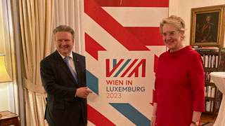 Herr im Anzug und Dame in rotem Kleid neben einem Roll-up mit Text "Wien in Luxemburg"