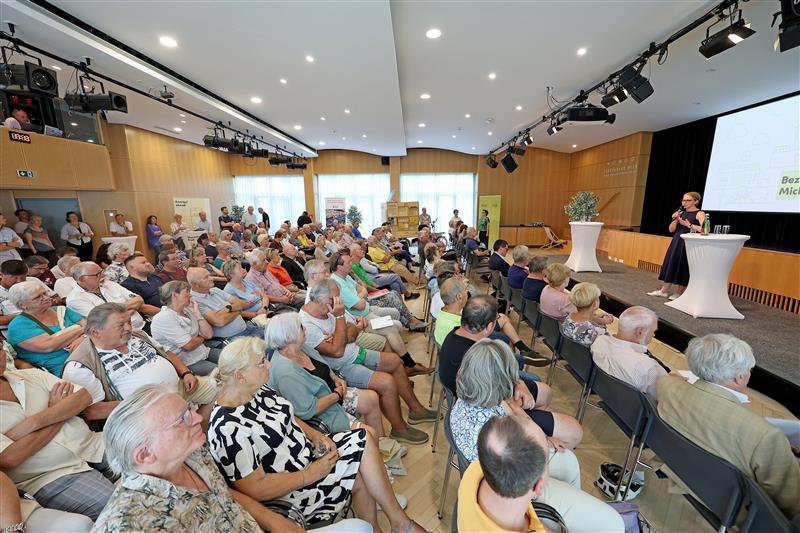 Großer Veranstaltungssaal mit vielen sitzenden Besucherinnen und Besucher, die einem Vortrag folgen. Eine Rednerin steht auf der Bühne vor einer Präsentation mit der Überschrift „Bezirk“. Copyright: Stadt Wien/Martin Votova
