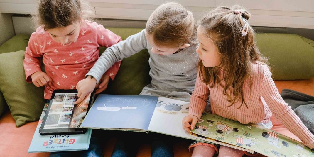 3 Kinder sitzen auf einem Sofa. Zwei Mädchen halten jeweils ein Buch. Das dritte Mädchen hält ein Tablet. Sie sind interessiert an den Informationen. 