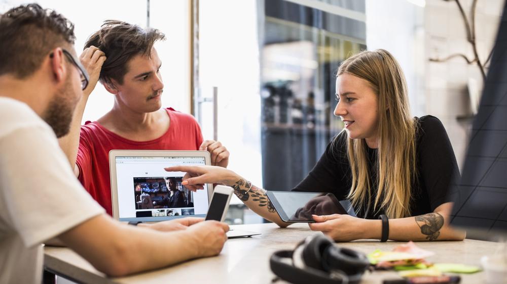zwei junge Männer und eine junge Frau sitzen auf einem Tisch. der Mann in der Mitte hat einen Laptop vor sich stehen und die junge Frau zeigt mit einem Finger auf den Bildschirm. 