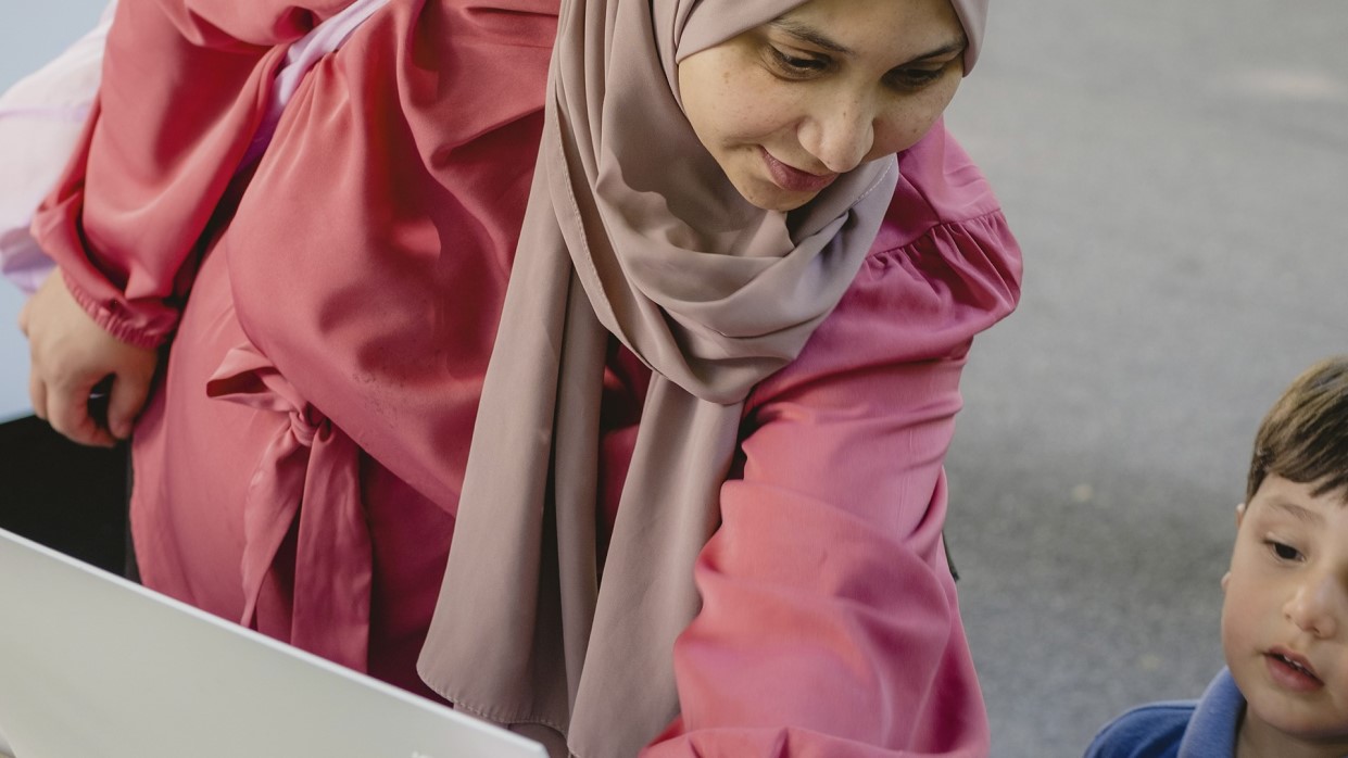 Kapitelbild: Eine Frau beugt sich über eine Bank mit weißen Schildern und zeigt einem Kind etwas darauf. Beide wirken aufmerksam. Die Szene findet im Freien statt.