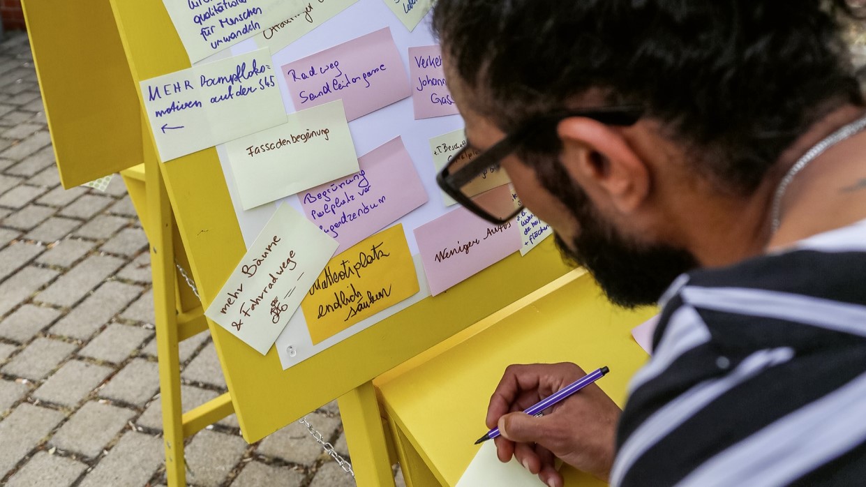 Kapitelbild: Eine Person schreibt eine Notiz auf einen farbigen Klebezettel. Vor ihr steht eine gelbe Pinnwand mit vielen bunten Notizen, auf denen Ideen und Wünsche für ein Wiener Quartier gesammelt sind.