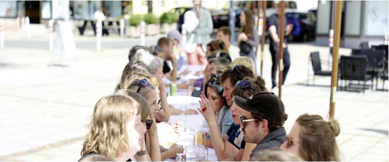 Titelbild: Viele Menschen an einem langen Tisch im Freien diskutieren