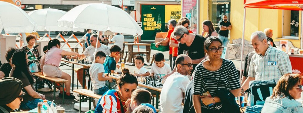 Menschen unterhalten sich auf Gastgartenbänken und Tischen im Freien – teilweise unter Sonnenschirmen.
