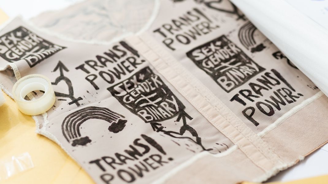 Textil, ein Oberteil?, auf einem Tisch mit den Aufschriften „SEX GENDER ist not BINARY“, „TRANS POWER!“, Am Rand ein Klebeband.
