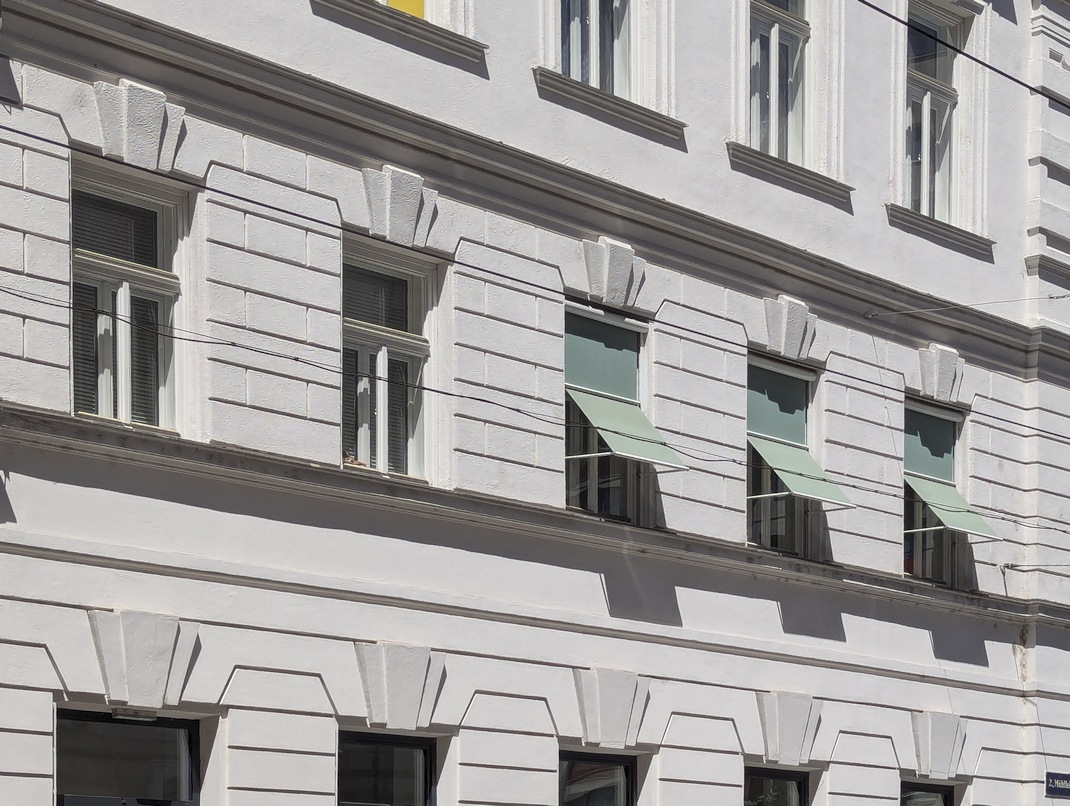 Markisoletten an einem dekorierten Gründerzeithaus in einer Schutzzone. Der runde Vorbaukasten findet in der Fensterlaibung der Kastenfenster Platz, die Konstruktion wird in Fensterfarbe ausgeführt. Der Textilbehang ist farbig. Die Sonnenschutzvorrichtungen an den einzelnen Fenstern des Hauses sind einheitlich nach diesem Prinzip gestaltet. 