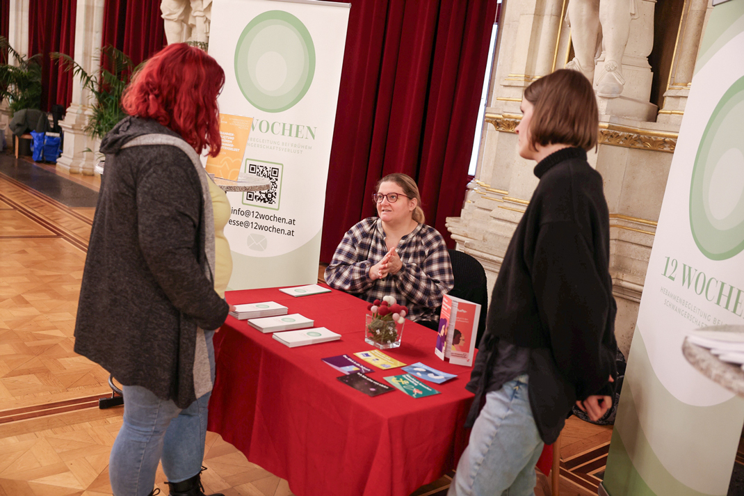 Zu sehen ist ein Tisch, an dem Infomaterialien aufliegen und eine Person sitzt, die gerade mit zwei anderen spricht.