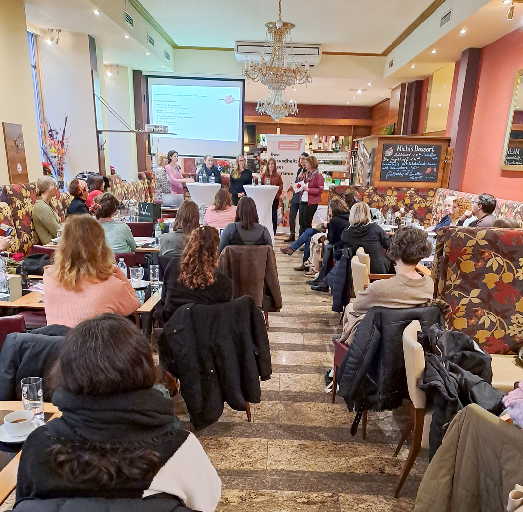 Zu sehen ist die Veranstaltung im Zuge des Podiumsgesprächs in einem Kaffeehaus. Die zahlreichen Besucher*innen schauen in Richtung der Vortragenden.
