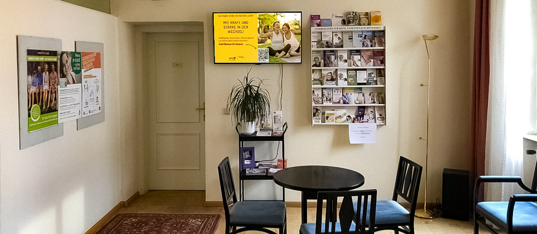 Blick in ein Wartezimmer. Am Bildschirm an der Wand läuft ein TV-Clip zum Thema Wechseljahre.