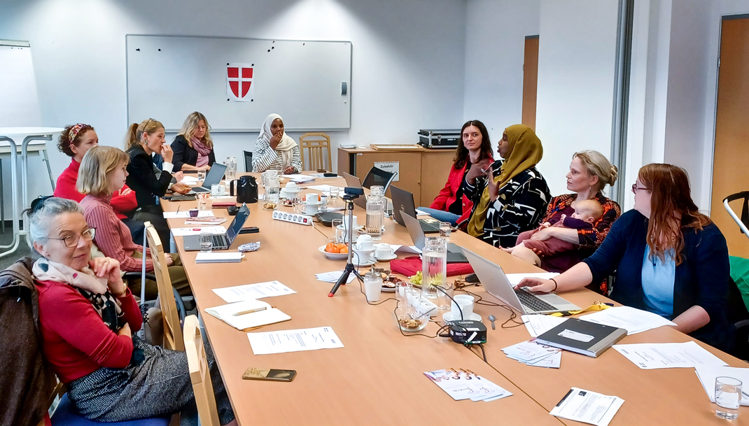 Zu sehen sind zirka zehn Frauen, die rund  um einen Tisch sitzen und gemeinsam diskutieren. Zetteln, Laptops und Gläser stehen auf dem Tisch. Eine Frau hät ein Baby in ihrer Schoß.