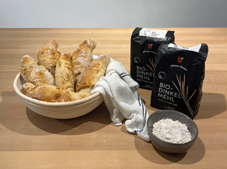 The photo shows freshly baked bread in a bowl on a wooden table. Next to it are a bowl of flour and two bags of organic spelt flour.