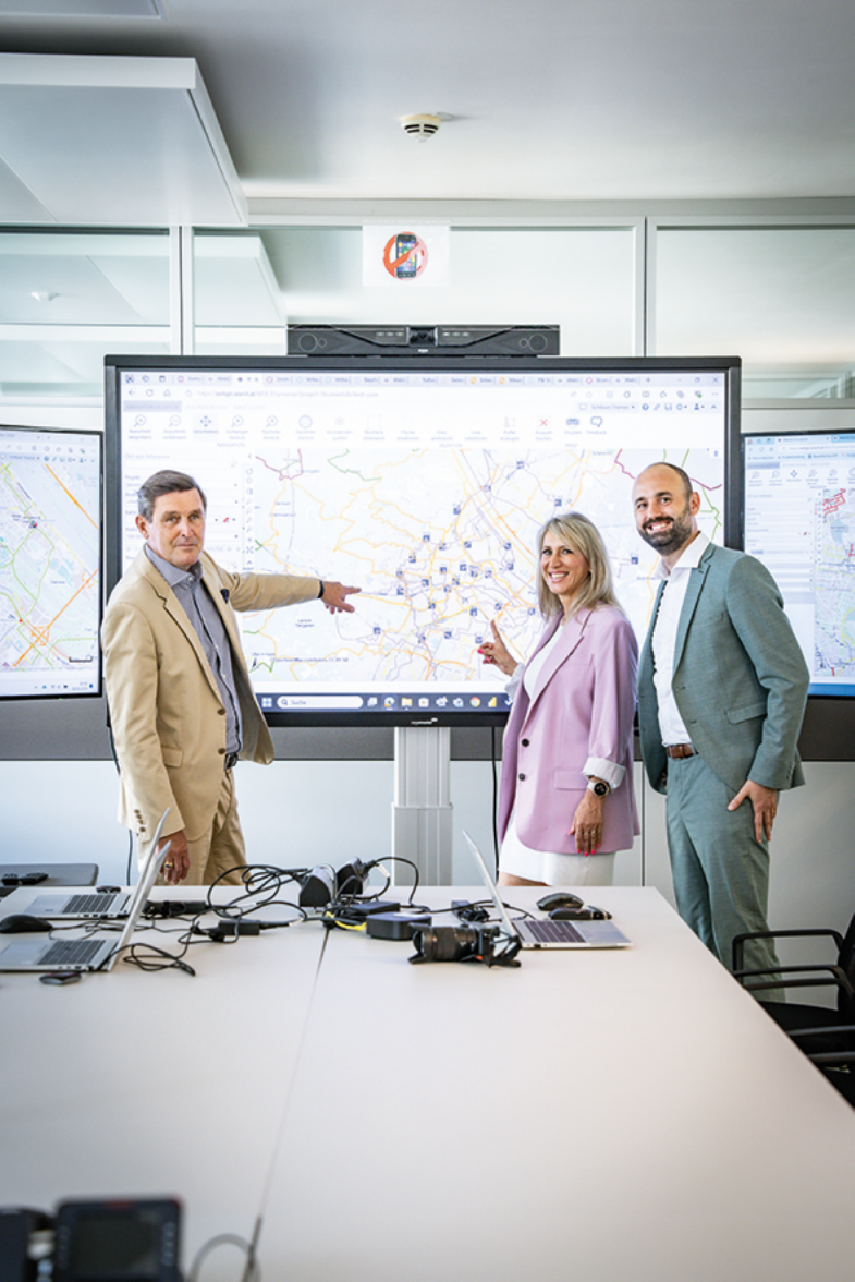 The photo shows two people standing in front of a large screen and smiling at the camera. The screen displays a map of Vienna.