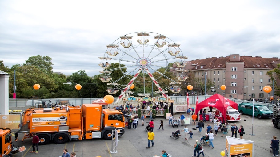 Kapitelbild: Auf einem offenen Platz steht ein großes Riesenrad; links stehen mehrere orangefarbene Nutzfahrzeuge. Zahlreiche Besucher*innen, darunter Eltern mit Kinderwägen, bewegen sich zwischen Informationsständen und Ständen auf dem Veranstaltungsgelände. (Copyright: Matern)