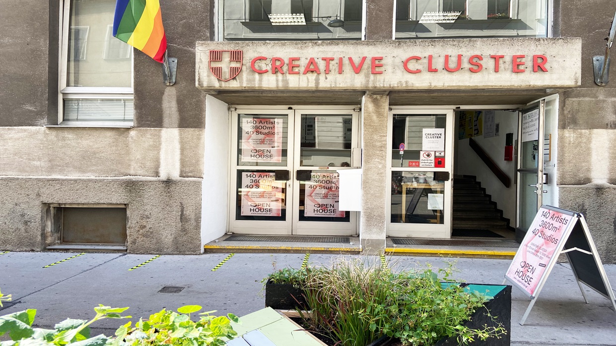 Kapitelbild: Das Foto zeigt den Eingangsbereich eines Gebäudes mit roter Beschriftung „CREATIVE CLUSTER“ über den Türen; Eine Regenbogenfahne hängt links. Vor dem Eingang stehen Pflanzenkästen. (Copyright: Katharina Egg)