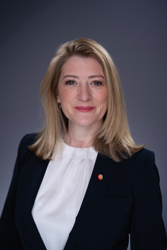 Formelles Porträt von Vizebürgermeisterin Mag. Bettina Emmerling: Sie trägt einen dunklen Blazer über einer weißen Bluse und hat schulterlange, hellblonde Haare. Der Hintergrund ist neutral-grau.