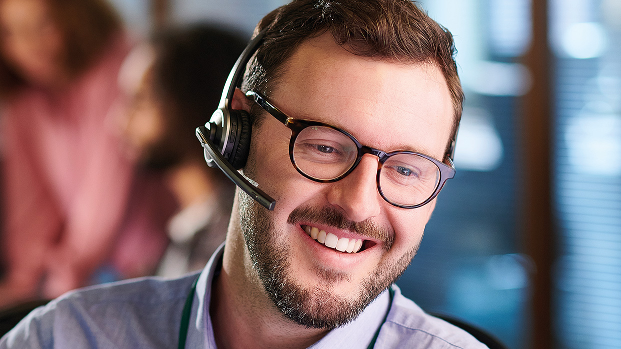 Kapitelbild: Porträtfoto eines lächelnden Mannes mit Brille und Headset, der offenbar in einem Büro arbeitet. Fokus liegt auf freundlicher Kundenbetreuung oder Support-Tätigkeit in einer professionellen Umgebung.