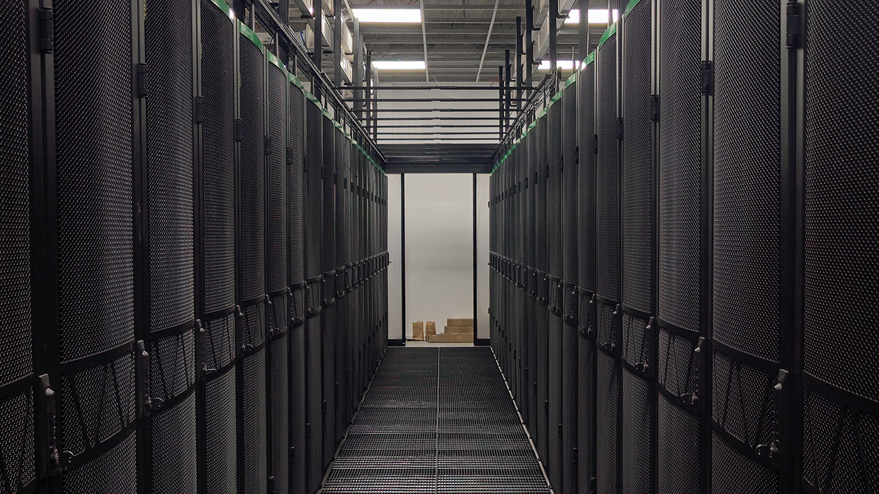 Kapitelbild: Fotografie eines Rechenzentrums mit zwei Reihen von schwarzen Server-Racks, die einen Gang in der Mitte bilden. Beleuchtung von der Decke beleuchtet den Gang, im Hintergrund sind einige Kartons zu sehen.