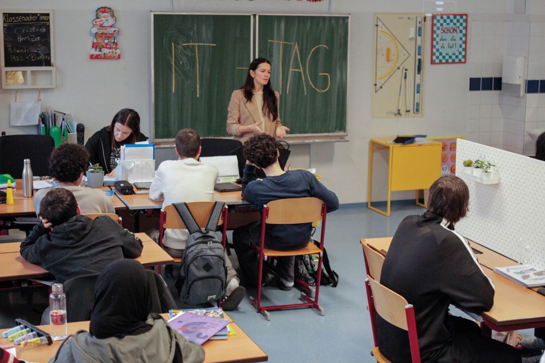 Fotografie eines Klassenzimmers mit IT-Expertin vor Tafel, auf der "IT-TAG" steht, und Schülern an Tischen mit Laptops und Schulmaterialien.