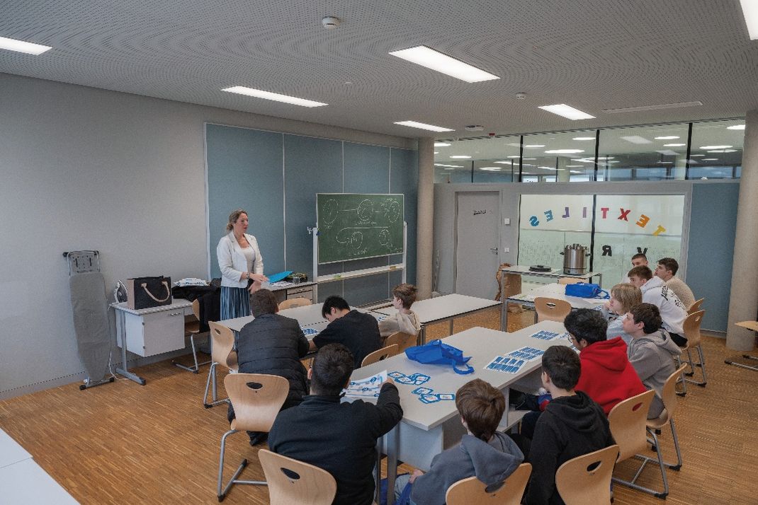 Fotografie eines Klassenzimmers mit einer Vortragenden, die vor einer Tafel steht und Schüler anspricht, die an Tischen sitzen. Raum hat helle Wände, Holzfußboden, Fenster mit Blick auf einen Flur und bunte Buchstaben an der Scheibe.