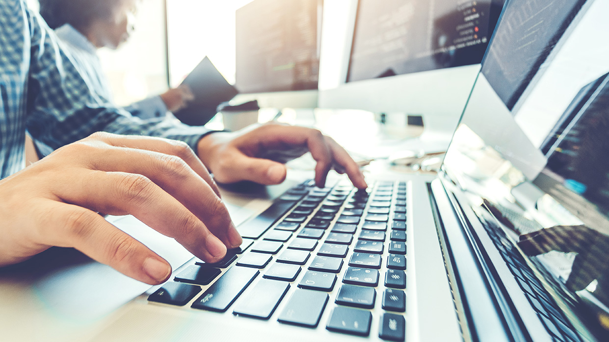 Kapitelbild: Fotografie zeigt Nahaufnahme von Händen, die auf Tastatur eines Laptops tippen, mit mehreren Monitoren im Hintergrund, die Programmiercode anzeigen. Kontext deutet auf Softwareentwicklung oder Programmierung hin, betont technische Arbeitsumgebung und Konzentration auf Codierung.