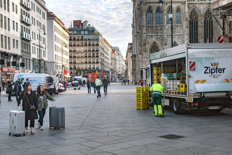 Am Stephansplatz in Wien wird ein Lastwagen entleert. Über den Platz gehen verschiedene Menschen, zwei Personen im Vordergrund haben einen Koffer.