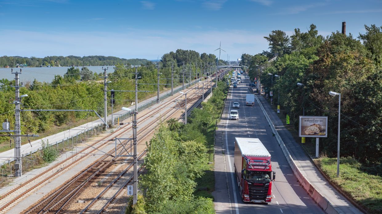 Kapitelbild: Neben einer Straße, auf der ein LKW fährt, sind Gleise zu sehen. Sowohl Gleise, als auch die Straße sind zwischen Alleen von Bäumen. Daneben fließt noch ein Fluss. 