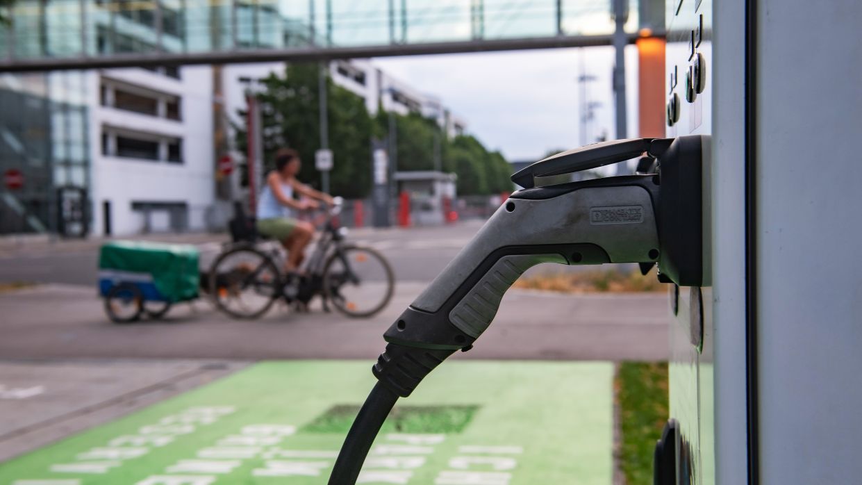 Kapitelbild: Eine elektrische Ladestation ist im Vordergrund, im Hintergrund fährt eine Person auf einem Fahrrad mit Anhänger. 

