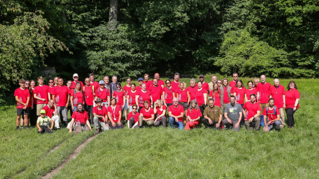 Expert*innen-Gruppen-Foto am Tag der Artenvielfalt 2022. Großteils rot gekleidete Menschen auf Wiese, dahinter Wald
