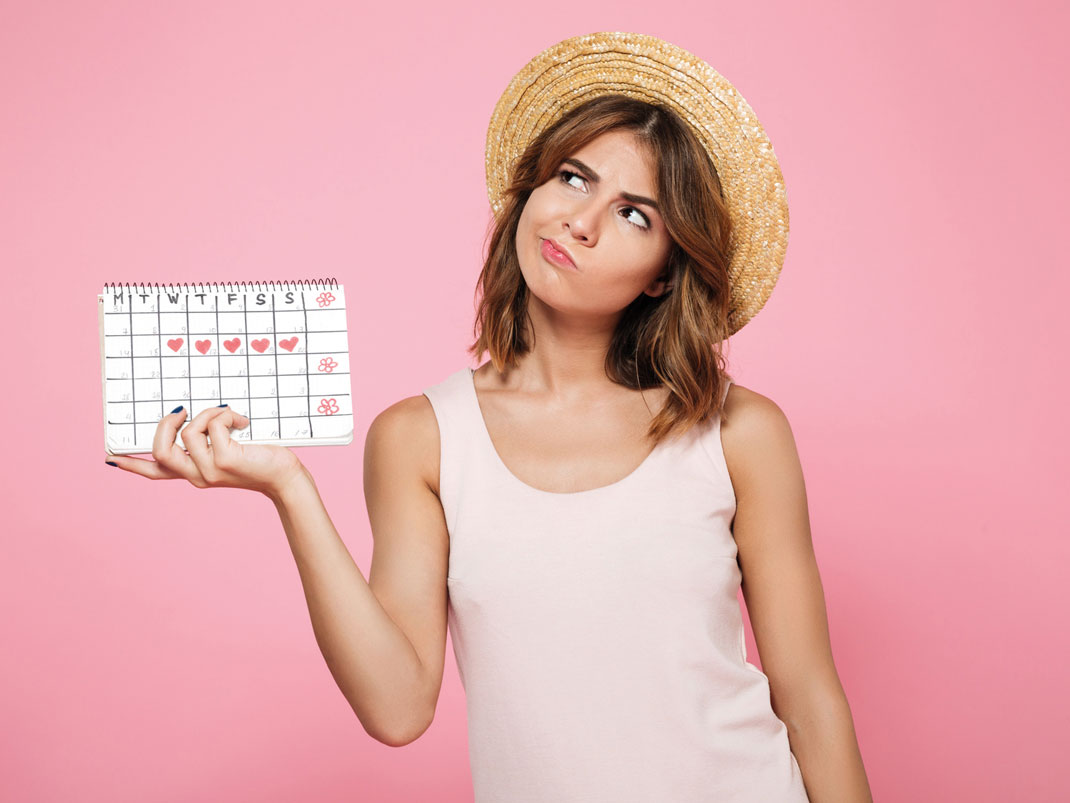 Fotografie einer Frau vor rosa Hintergrund, die einen Wochen-Kalender in der Hand hält. 5 Wochentage sind mit markierten Herzsymbolen gekennzeichnet.
