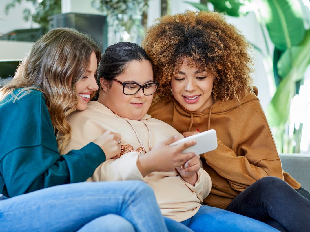 Fotografie von drei jungen Frauen, die eng nebeneinandersitzen und gemeinsam auf ein Smartphone schauen.