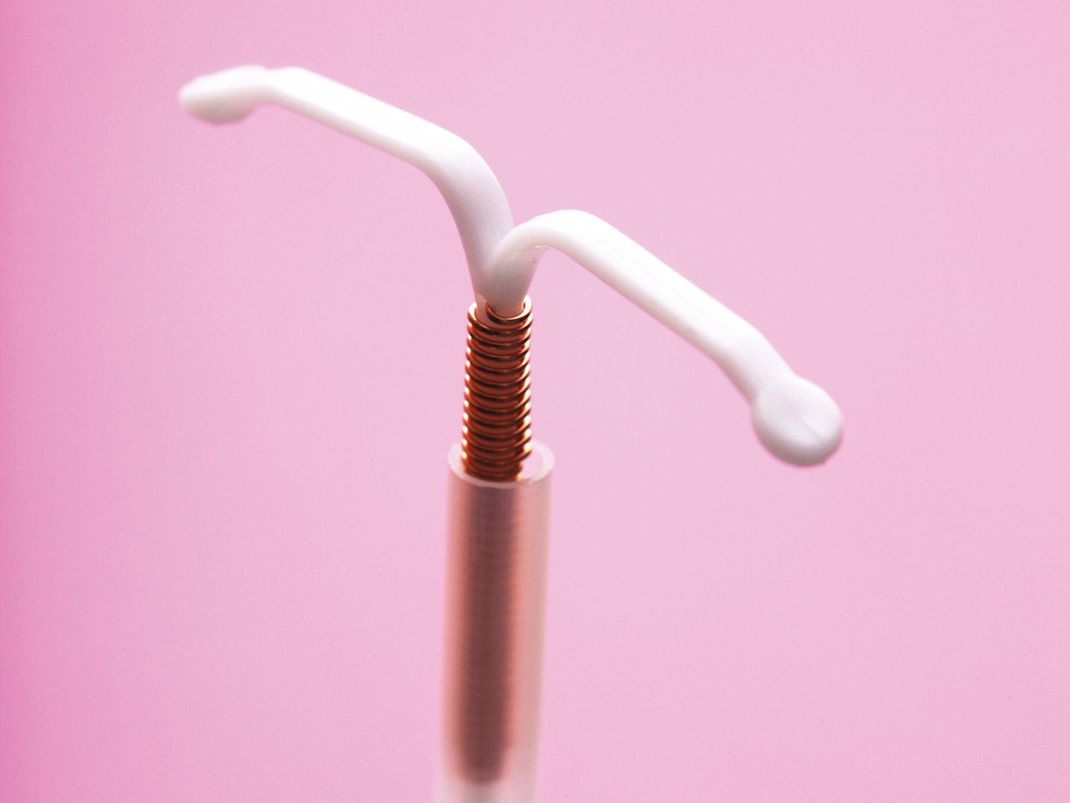 Nahaufnahme eines Intrauterinpessars (IUP) mit T-förmigem weißen Kunststoffkörper und spiralförmiger Kupferwicklung. Das Verhütungsmittel steht vor einfarbigem rosa Hintergrund.