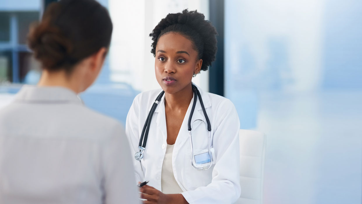 Kapitelbild: Zwei Frauen im Gespräch, eine trägt weißen Kittel und Stethoskop und hört einer nur von hinten sichtbaren Frau zu.  Es veranschaulicht die Kommunikation zwischen Patientin und Ärztin. © istockphoto.com