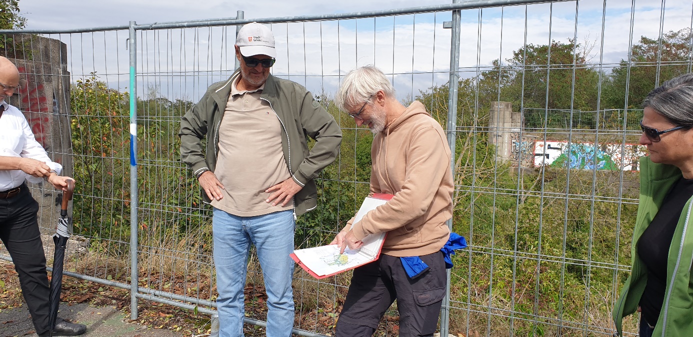 Kapitelbild: Mehrere Personen stehen an einem Metallzaun. Eine Person zeigt auf einen roten Ordner mit einem Plan. Die anderen hören zu. Im Hintergrund eine verwilderte Grünfläche.
@ Uschi Dorau,. UIV
