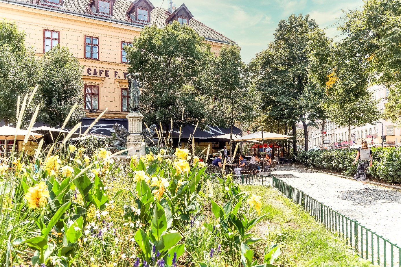 Niegrig umzäunte Grünfläche mit Gräsern und Blumen, im Hintergrund ein Café mit Sonnenschirmen und Leuten darunter.