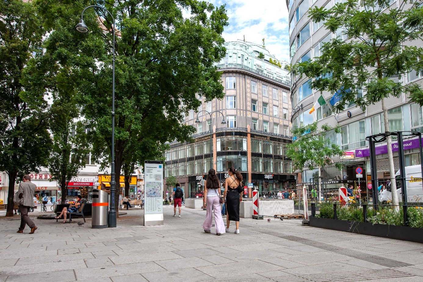 Zwei Frauen in einer Fußgängerzone in einem zentralen Viertel. Baume, weitere Fußgänger und Gebäude weiter im Hintergrund.