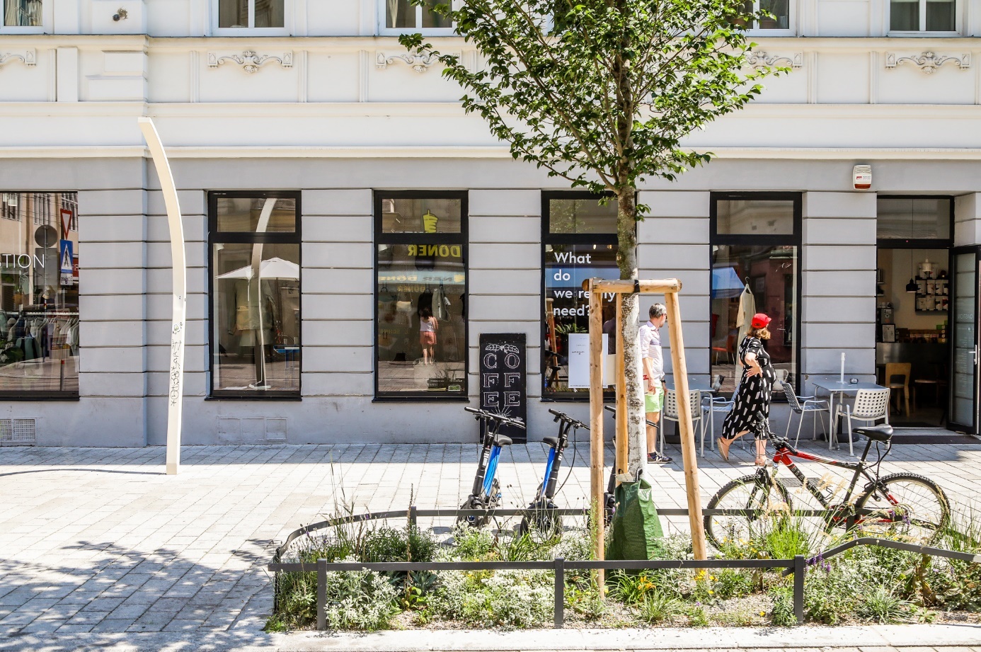 Ein Bild, das draußen, Fahrradreifen, Gebäude, Rad enthält.

Gesteig mit Grüninsel in der ein junger Baum mit Holzstütze wächst. Dahinter Roller, ein Fahrrad, zwei Leute und Geschäfte.