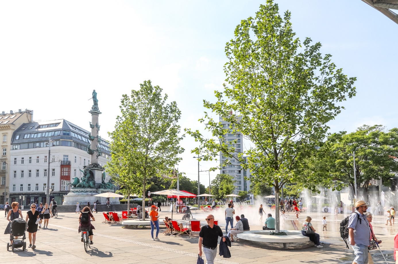Fußgängerbereich am Praterstern mit Menschen im Sonnenlicht, aktivierten Wasserspielen, Bäumen, Säule und Gebäuden.