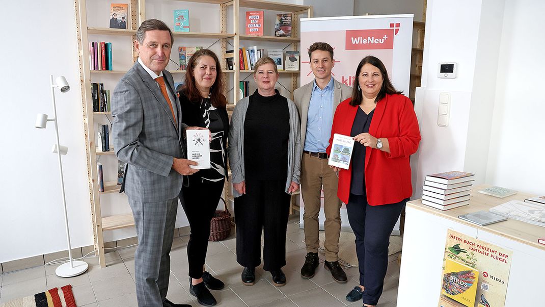 Five people standing in a row, looking at the camera in front of a bookshelf, two of whom are holding books up towards the camera.