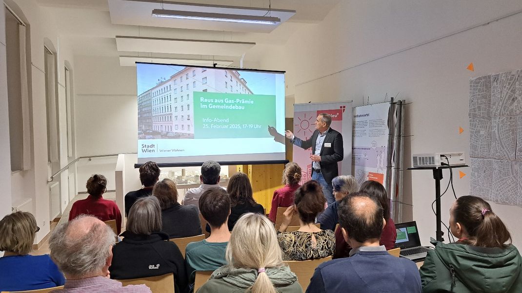 Audience members at the back; in front of them, in the background of the picture, a person giving a talk next to a screen showing a Gründerzeit-era building and text overlays.