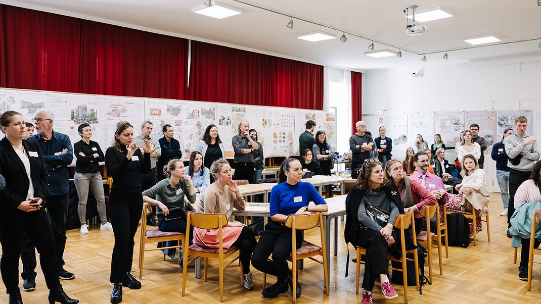 A large number of people, some sitting at tables and others standing, looking towards a stage. In the background, on the wall, are rows of project posters created by students.
